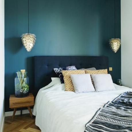 Tyylikkäässä makuuhuoneen sisustuksessa pieni puinen yöpöytä, puutarha purkissa, valkoiset vuodevaatteet, väripeitset ja viltti. Tila sinisillä seinillä ja ruskealla puisella parketilla. Suunnittelulamppu.