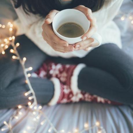 nainen istuu kotona jalat ristissä sängyssä, jolla on kuppi kahvia, joulukoristeita